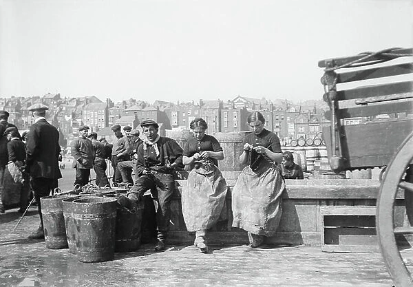Female herring fishers, knitting on the dock, in front of empty buckets waiting for the return of fishing, in Scarborough (Yorkshire, England), 1910 (b / w photo)