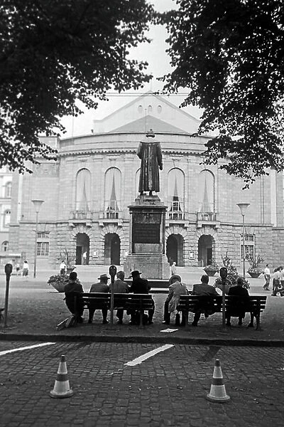 Inhabitants of Mainz sitting on benches on Gutenbergplatz square, facing the rear of the Gutenberg monument and the city theatre, Germany 1961