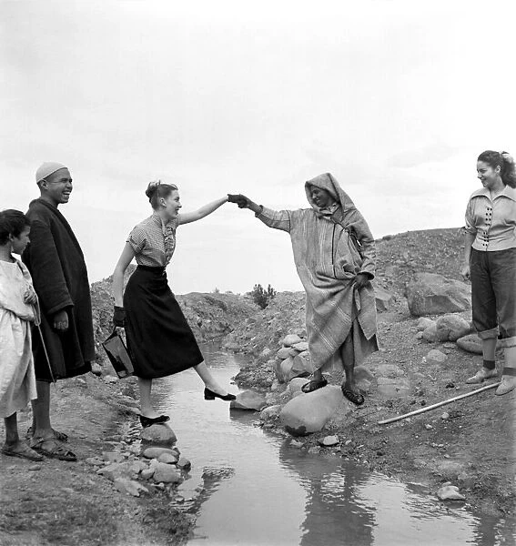 Man in arabic dress lends his hand to help French actresses across a stream as he guides