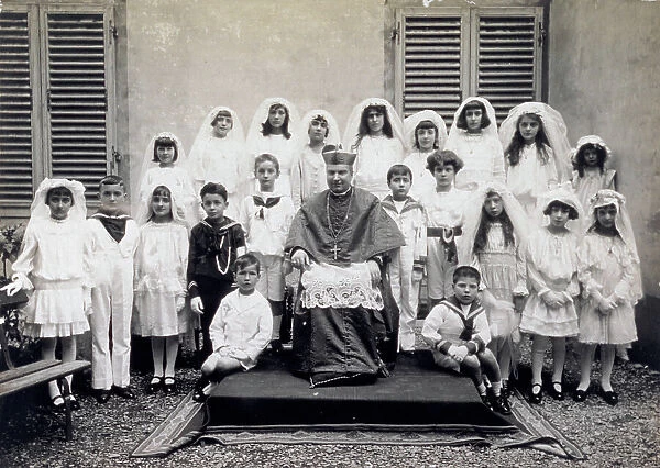 Group of children, in first communion attire, shown with the bishop. The boys wear sailor outfits and the little girls wear veils