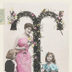 T: Capital letter decorated with flowers, a woman wearing a long cloth and a boy and a girl holding a basket of flowers. 1907 (photograph)